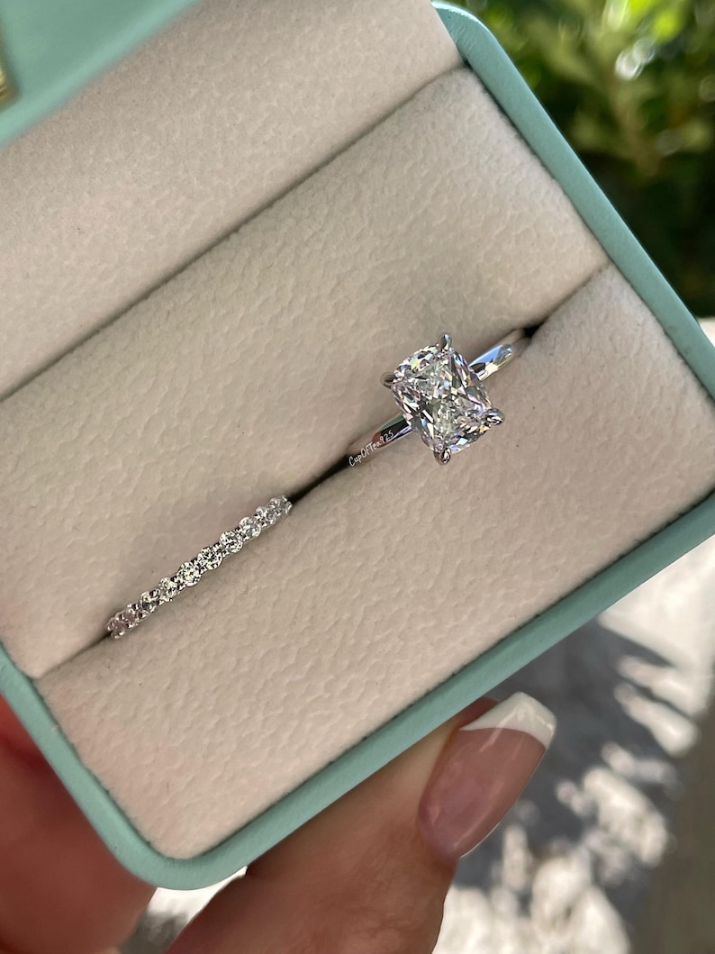 May include: A silver-toned engagement ring set featuring a cushion-cut center stone on a thin band and a matching eternity band with small round stones. The rings are displayed in a light blue box with a beige interior lining.