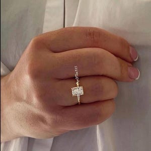 May include: A gold ring with a large, clear, square-shaped gemstone. The ring is on a finger.