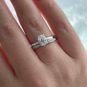 May include: A silver oval engagement ring with a large center stone and a smaller band with pave diamonds. The ring is on a finger with a light pink nail polish.