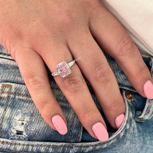 Peut inclure: Une bague en argent avec un gros diamant rose de forme carrée. La bague est portée sur un doigt, qui repose sur un jean bleu.