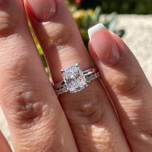 May include: A silver-toned ring set featuring a large, rectangular cushion-cut center stone held by four prongs on a smooth band, paired with a matching eternity band adorned with a row of small, round, brilliant-cut stones.