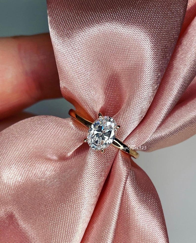 Puede incluir: Un anillo de oro con una gran piedra preciosa ovalada transparente. El anillo est&aacute; sobre una tela de sat&eacute;n rosa.