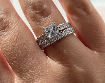 Puede incluir: Un anillo de plata con una gran piedra de circonita cúbica de corte cuadrado rodeada de piedras más pequeñas. El anillo se lleva en el dedo anular de una mano.
