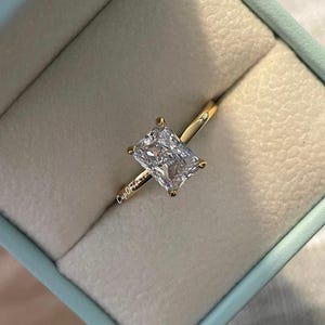 May include: A gold ring with a large, clear, rectangular-shaped diamond in a ring box. The ring box is light blue.