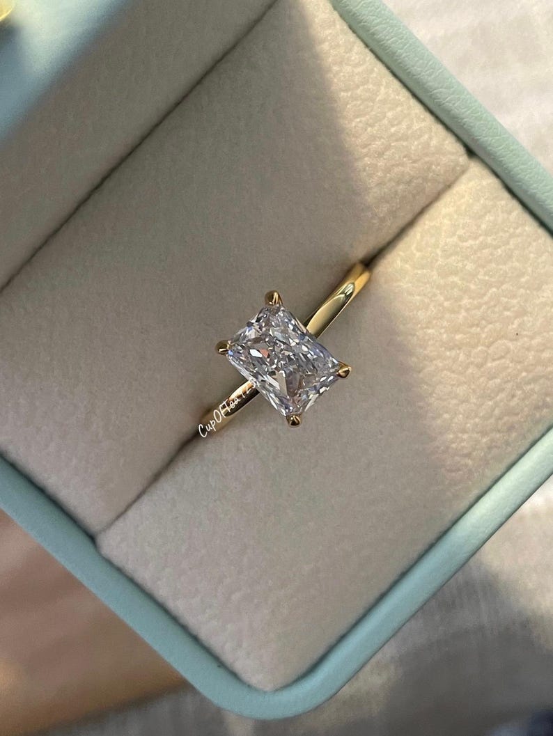 May include: A gold ring with a large, clear, rectangular-shaped diamond in a ring box. The ring box is light blue.