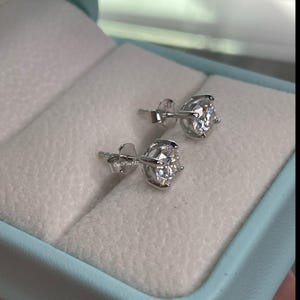 May include: A pair of silver stud earrings with clear round stones. The earrings are in a light blue box with white lining.