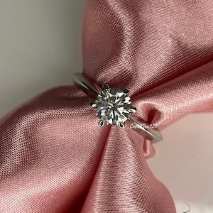 May include: A silver ring with a large, clear, round diamond set in a prong setting. The ring is on a pink satin fabric.