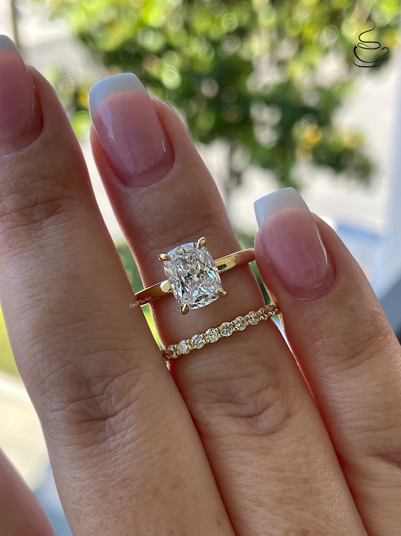 May include: A gold ring set consisting of a solitaire ring with a cushion-cut clear stone held by four prongs and a matching thin band featuring a row of small round clear stones. Both rings are crafted from polished yellow gold metal.