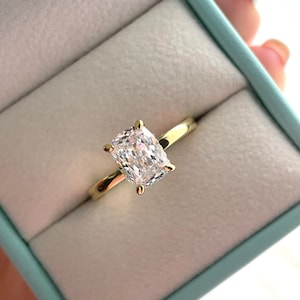 May include: A gold ring featuring a single cushion-cut clear gemstone set in a four-prong setting. The ring is presented inside a light blue jewelry box with a soft white interior lining, showcasing the polished metal band and faceted stone.