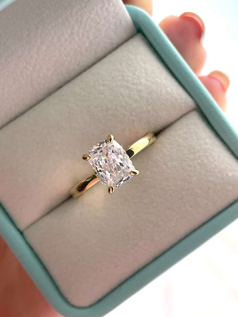 May include: A gold ring featuring a single cushion-cut clear gemstone set in a four-prong setting. The ring is presented inside a light blue jewelry box with a soft white interior lining, showcasing the polished metal band and faceted stone.