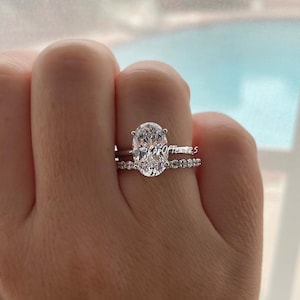 May include: A silver engagement ring with a large oval-shaped diamond and a matching band with smaller diamonds. The ring is on a person's finger.