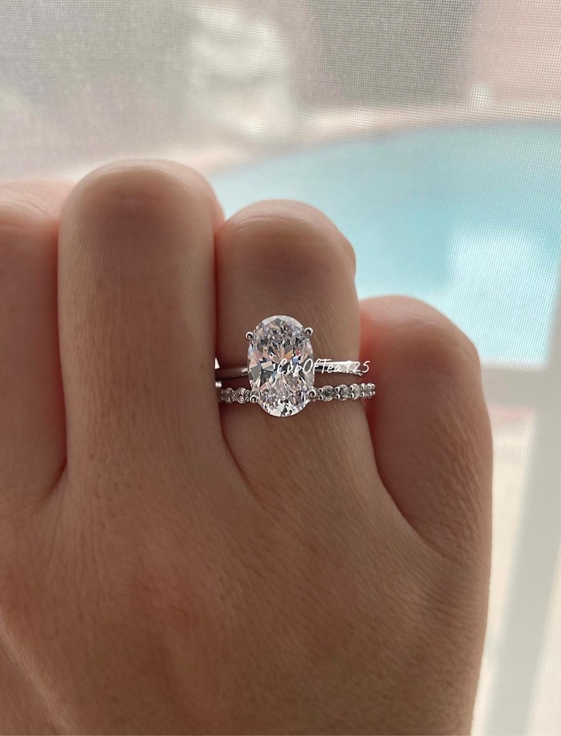 May include: A silver engagement ring with a large oval-shaped diamond and a matching band with smaller diamonds. The ring is on a person's finger.