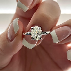 May include: A silver ring with a large oval-shaped clear diamond. The ring is on a finger with a white french manicure.
