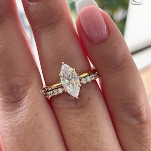 May include: A gold engagement ring with a large pear-shaped diamond and a smaller diamond band. The ring is on a finger with a light pink nail polish.