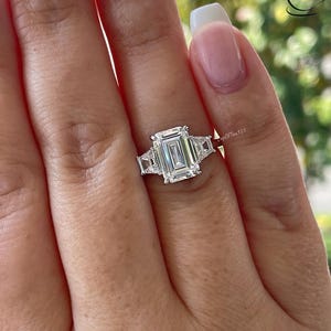 Pode incluir: Um anel de prata com uma grande pedra central de diamante de corte esmeralda e duas pedras de diamante de acento menores de cada lado.