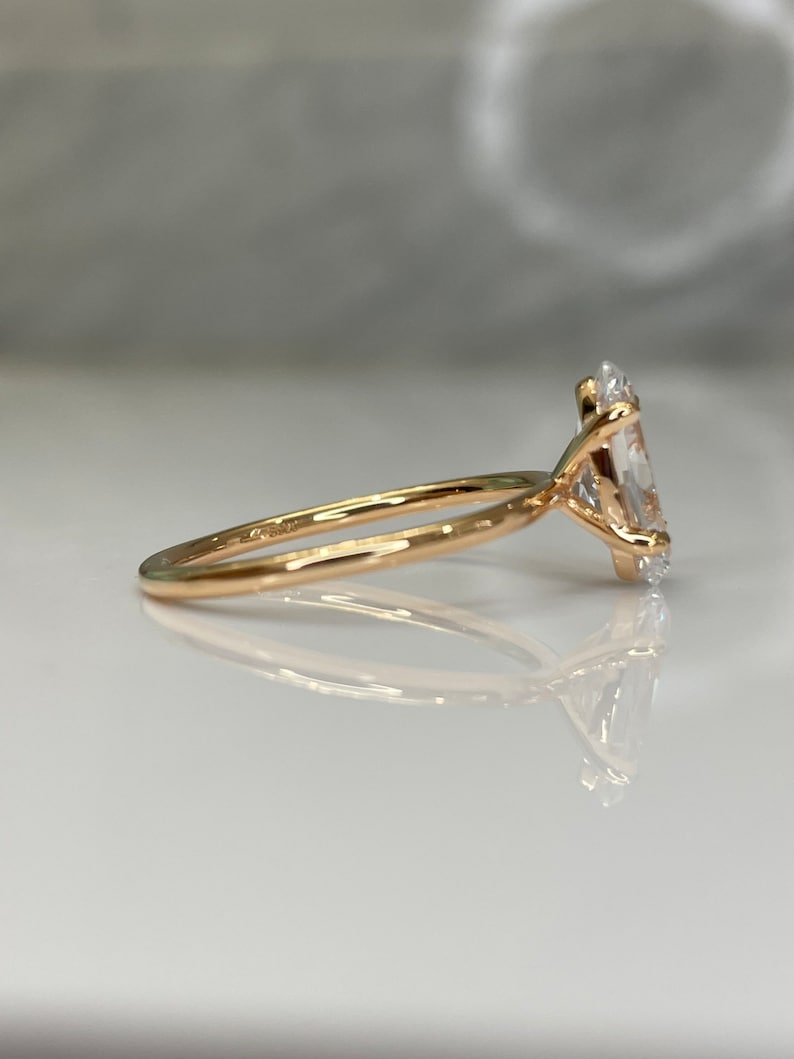 Puede incluir: Un anillo de compromiso de oro con un gran diamante ovalado transparente. El anillo est&aacute; engastado en una banda fina.