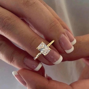 May include: A gold ring with a large, rectangular, clear gemstone. The ring is on a finger with a white french manicure.