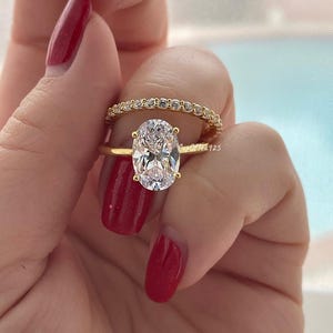 May include: A gold ring set with a large oval-shaped clear gemstone. The ring is paired with a thin gold band set with small clear gemstones.