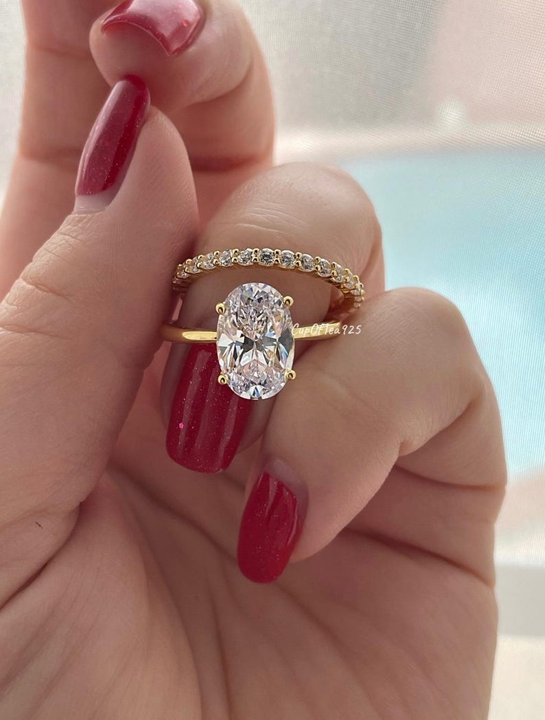 May include: A gold ring set with a large oval-shaped clear gemstone. The ring is paired with a thin gold band set with small clear gemstones.
