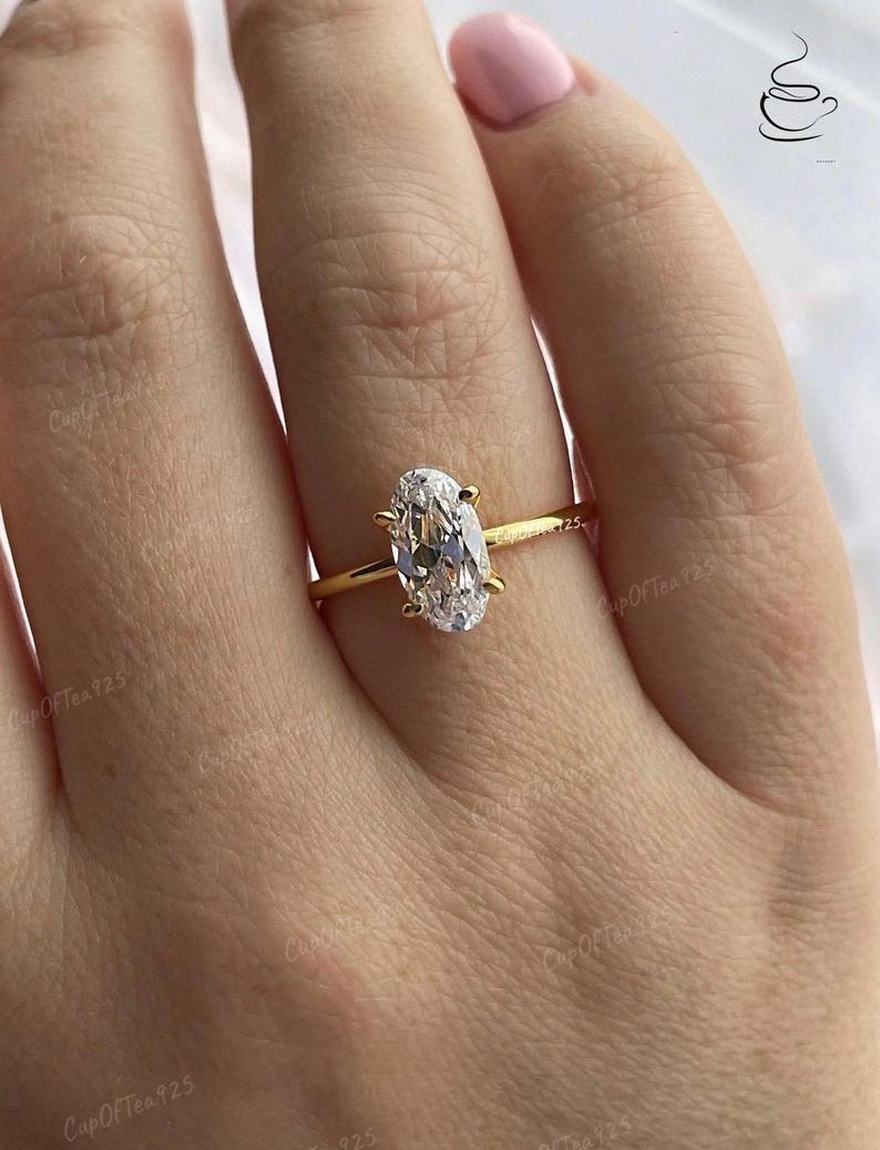 Puede incluir: Un anillo de oro con una gran piedra preciosa ovalada transparente. El anillo est&aacute; en el dedo de una persona.