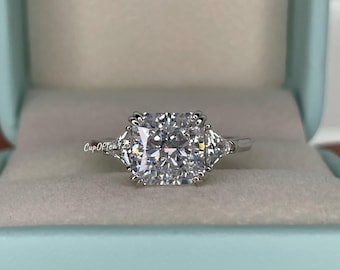 Pode incluir: Um anel de prata com um grande diamante quadrado no centro e dois diamantes triangulares menores de cada lado. O anel está em uma caixa azul claro.