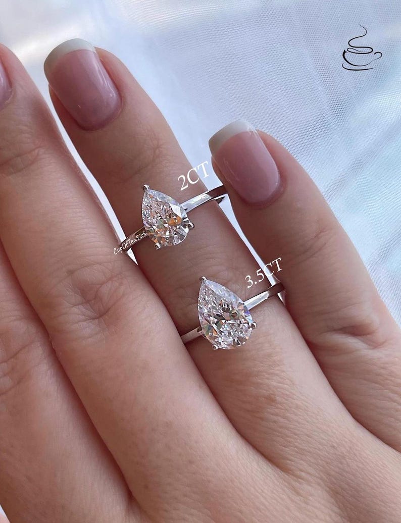 K&ouml;nnte beinhalten: Zwei birnenf&ouml;rmige Diamant-Verlobungsringe an einer Hand. Der Ring links hat einen 2 Karat Diamanten und der Ring rechts einen 3,5 Karat Diamanten.