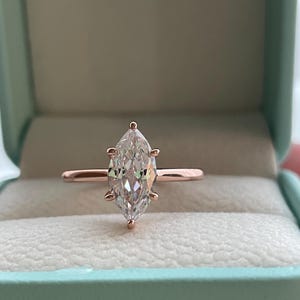K&ouml;nnte beinhalten: Ein Verlobungsring aus Ros&eacute;gold mit einem Diamanten im Marquiseschliff. Der Diamant ist mit vier Krappen gefasst und wird in einer hellblauen Ringschatulle pr&auml;sentiert. Der Ring hat ein schlankes, poliertes Band.