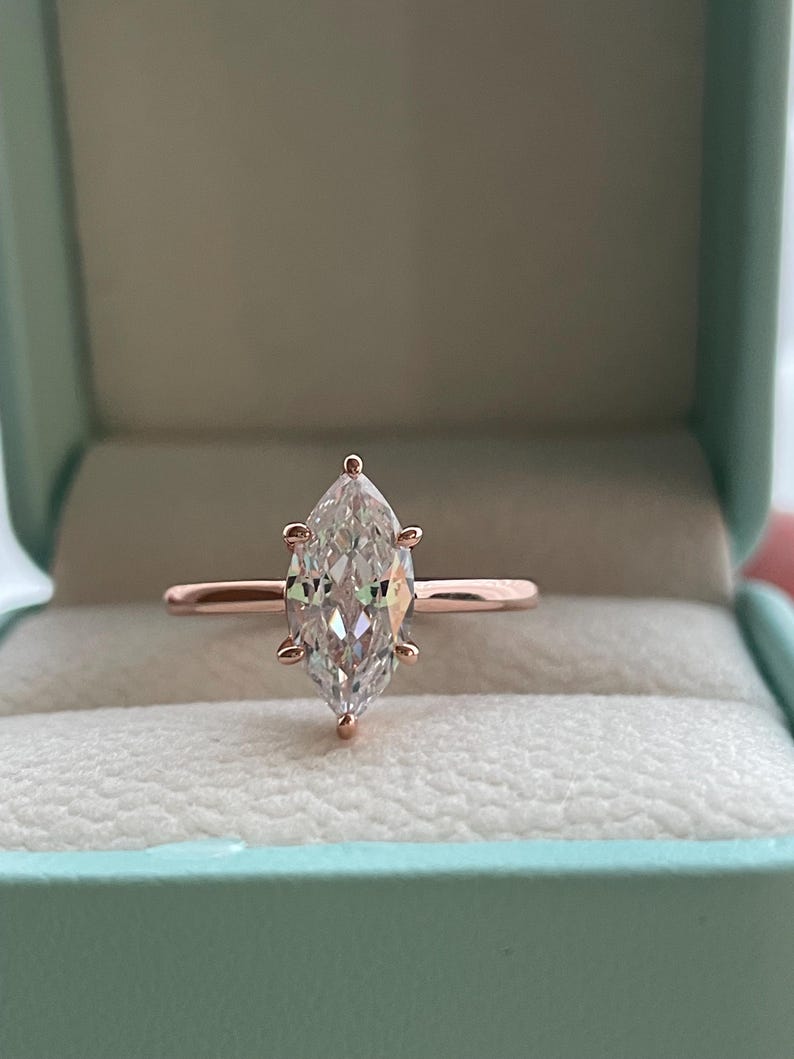 K&ouml;nnte beinhalten: Ein Verlobungsring aus Ros&eacute;gold mit einem Diamanten im Marquiseschliff. Der Diamant ist mit vier Krappen gefasst und wird in einer hellblauen Ringschatulle pr&auml;sentiert. Der Ring hat ein schlankes, poliertes Band.