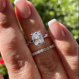 May include: A silver-toned ring set featuring a large cushion-cut center stone held by four prongs on a plain band, paired with a matching eternity band encrusted with a row of small, round, brilliant-cut stones that sparkle in the sunlight.