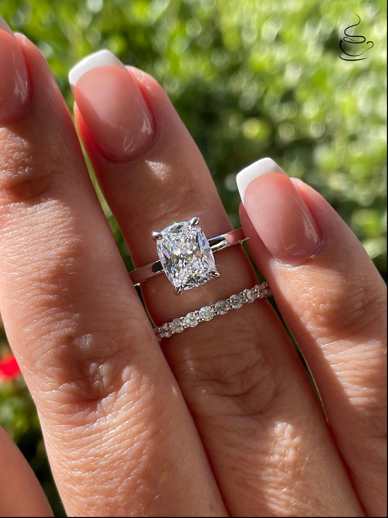 May include: A silver-toned ring set featuring a large cushion-cut center stone held by four prongs on a plain band, paired with a matching eternity band encrusted with a row of small, round, brilliant-cut stones that sparkle in the sunlight.