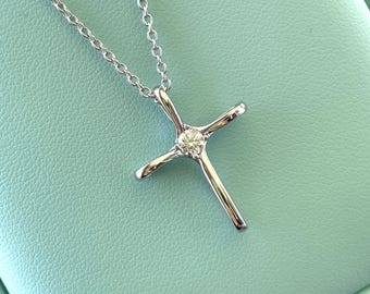 May include: A silver-colored cross pendant necklace with a small, round, clear gemstone at the center. The cross is suspended from a delicate chain. The background is a light teal color.