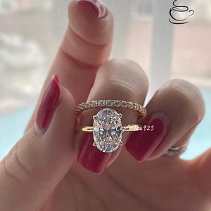 May include: A gold ring set with a large oval-shaped clear gemstone. The ring is worn on a finger with red nail polish. A smaller band of gold with clear gemstones is worn on the same finger.