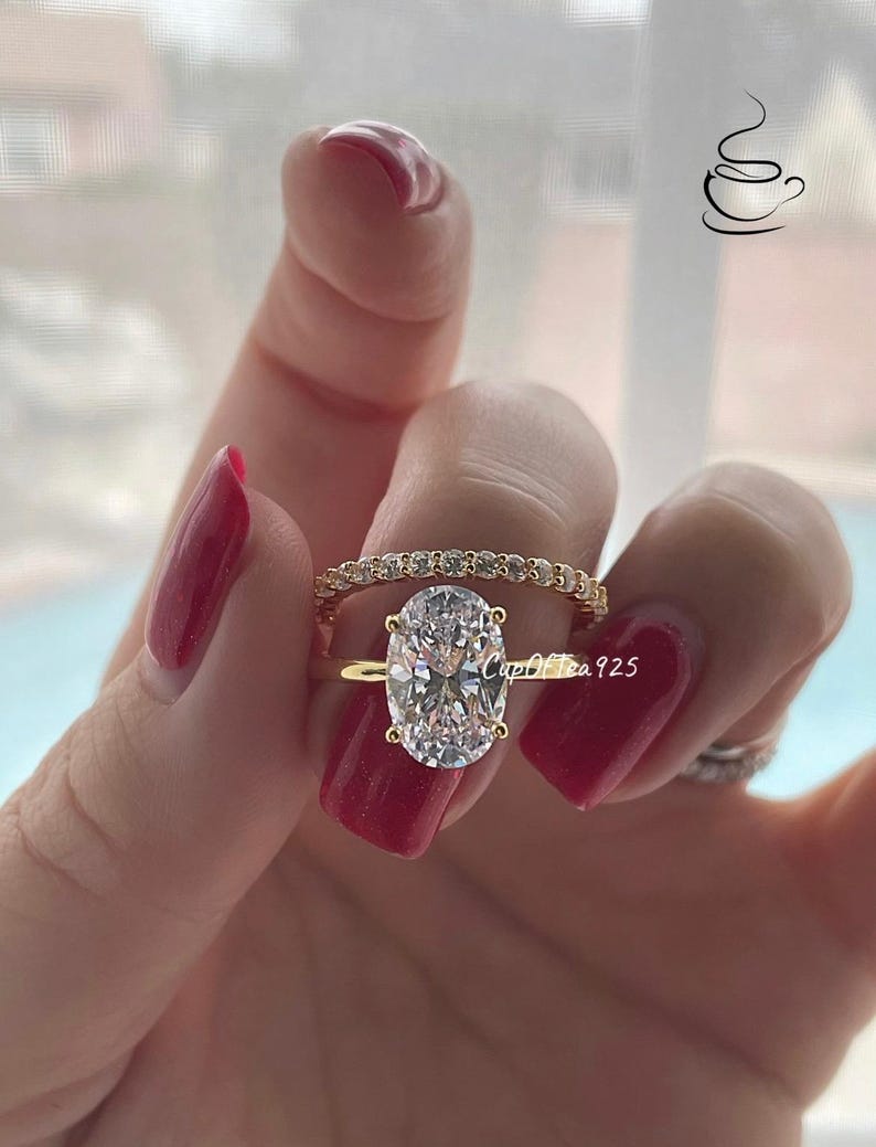 May include: A gold ring set with a large oval-shaped clear gemstone. The ring is worn on a finger with red nail polish. A smaller band of gold with clear gemstones is worn on the same finger.