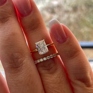 May include: A gold ring set consisting of a solitaire ring with a large rectangular radiant-cut clear stone and a matching thin band featuring a row of small round clear stones. The rings are worn together on a finger with a French manicure.