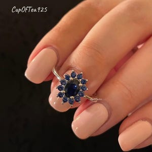May include: A silver ring with a large oval blue gemstone surrounded by smaller blue gemstones. The ring is on a finger with light pink nail polish.