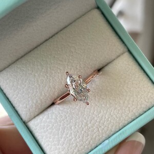 K&ouml;nnte beinhalten: Ein Verlobungsring aus Ros&eacute;gold mit einem Diamanten im Marquiseschliff, gefasst in einer sechs-Zacken-Fassung. Der Ring wird in einer hellblauen Ringschatulle mit wei&szlig;em Innenraum pr&auml;sentiert. Der Ring hat ein schmales Band.