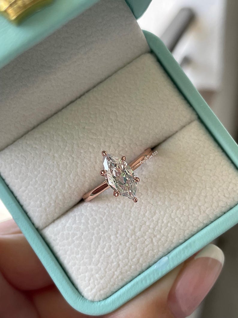 K&ouml;nnte beinhalten: Ein Verlobungsring aus Ros&eacute;gold mit einem Diamanten im Marquiseschliff, gefasst in einer sechs-Zacken-Fassung. Der Ring wird in einer hellblauen Ringschatulle mit wei&szlig;em Innenraum pr&auml;sentiert. Der Ring hat ein schmales Band.