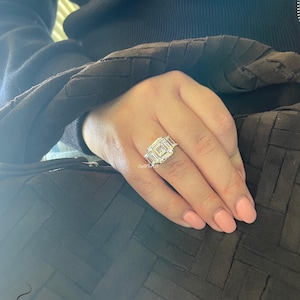 May include: A large, square-shaped diamond ring with smaller diamonds on the band. The ring is on a person's finger.