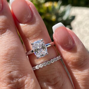 May include: A silver-toned engagement ring featuring a large cushion-cut center stone with four prongs, paired with a matching thin eternity band set with small round stones. The band of the engagement ring has the text CupOfTea125 engraved.