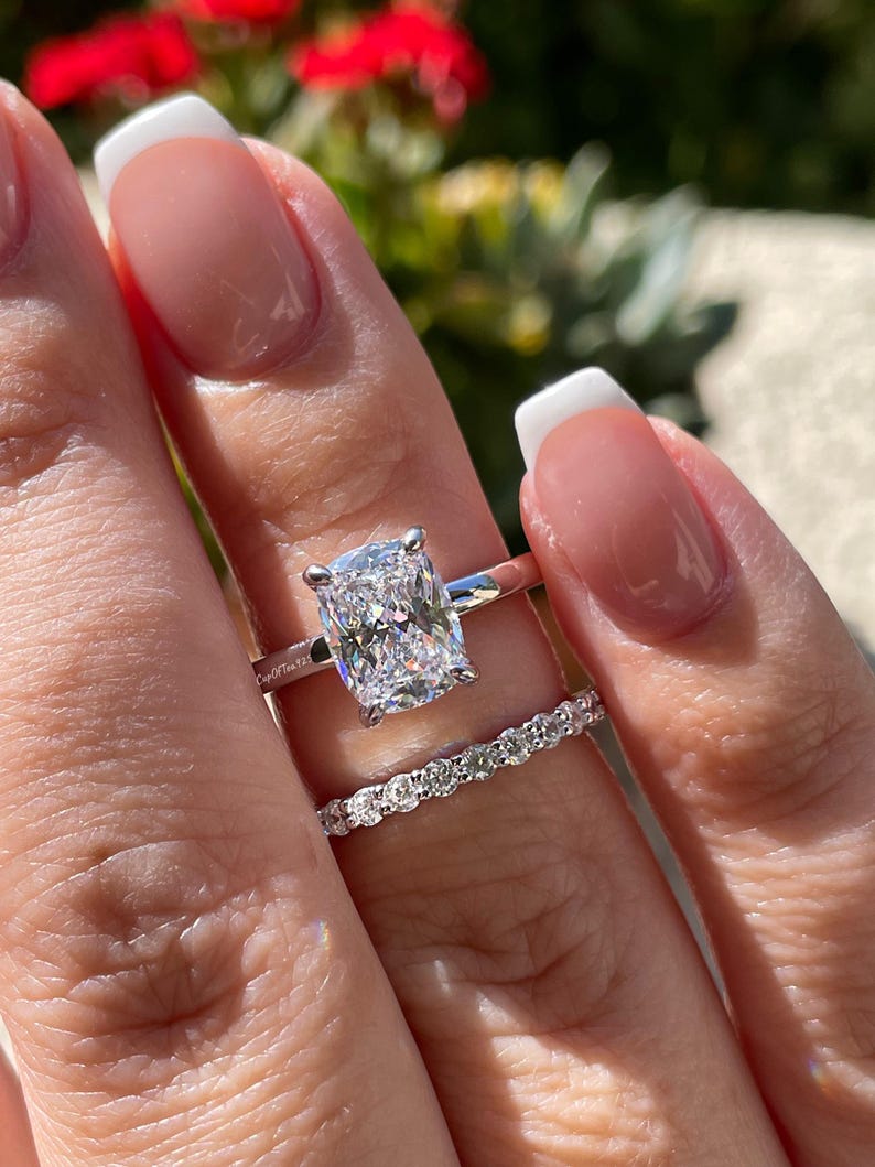 May include: A silver-toned engagement ring featuring a large cushion-cut center stone with four prongs, paired with a matching thin eternity band set with small round stones. The band of the engagement ring has the text CupOfTea125 engraved.