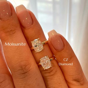 May include: Two gold rings featuring cushion-cut stones. The top ring is labeled Moissanite, and the bottom ring is labeled CZ Diamond. Both rings have a simple gold band and a four-prong setting holding the clear, faceted center stones in place.