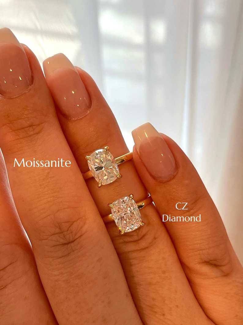 May include: Two gold rings featuring cushion-cut stones. The top ring is labeled Moissanite, and the bottom ring is labeled CZ Diamond. Both rings have a simple gold band and a four-prong setting holding the clear, faceted center stones in place.