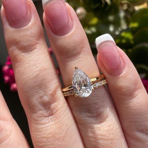 May include: A pear-shaped diamond engagement ring with a gold band and a smaller diamond band. The ring is set with a single large pear-shaped diamond in the center and a row of smaller diamonds on the band.