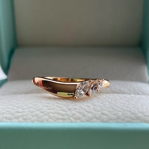 May include: A gold ring featuring three marquise-cut gemstones. The ring has a curved design and is set in a gold band. The ring is presented in a light blue box.
