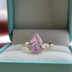 May include: A gold ring featuring a large, pear-shaped pink gemstone as the centerpiece, flanked by two smaller, clear gemstones. The ring is presented in a light blue jewelry box with a white interior.