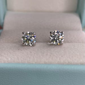 May include: A pair of diamond stud earrings in a light blue jewelry box. The earrings are round and have a clear, sparkling appearance.