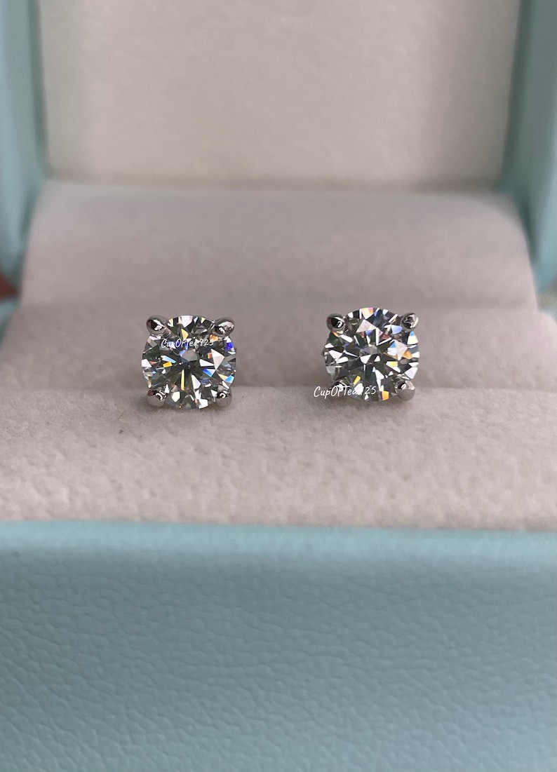 May include: A pair of diamond stud earrings in a light blue jewelry box. The earrings are round and have a clear, sparkling appearance.