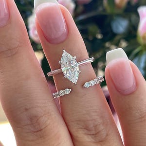 Może przedstawiać: Srebrny pierścionek z dużym diamentem w kształcie markizy i mniejszą obrączką z diamentów. Pierścionek jest na palcu z białym manicure.