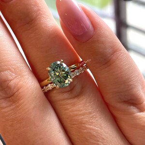 以下が含まれることがあります： 4本の爪で留められた中央のオーバルカットの緑色の宝石と、小さな透明な石が埋め込まれたマッチングバンドで構成されるゴールドのリングセット。バンドにはCupOfTea925の刻印があり、磨き上げられた金属光沢仕上げが施されています。