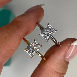 May include: Two engagement rings with square-shaped cubic zirconia stones. The ring on the left has a gold band, and the ring on the right has a silver band.